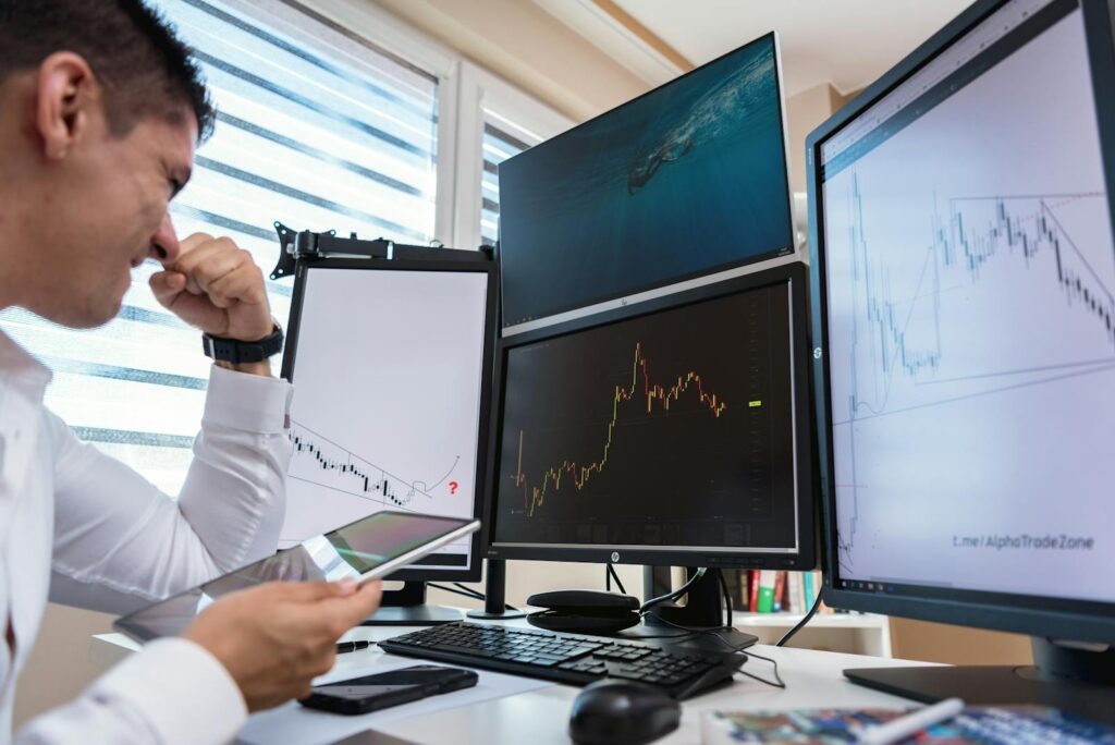 Trader in white shirt analyzing stock charts on multiple monitors during daytime in an office setting.