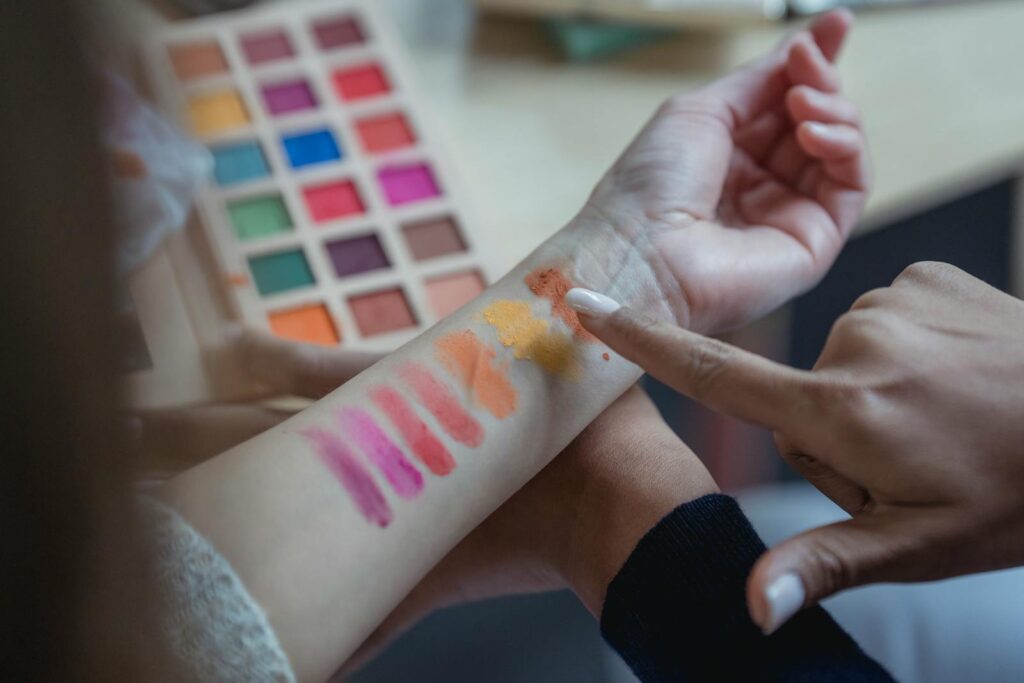 From above of crop unrecognizable young female makeup artist pointing at arm of client while applying colorful eyeshadows on wrist for testing